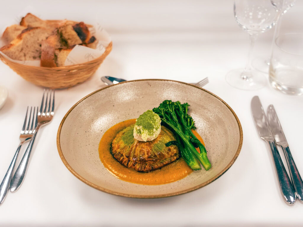 Deconstructed scallop with greens and vibrant orange sauce on elegant ceramic plate.